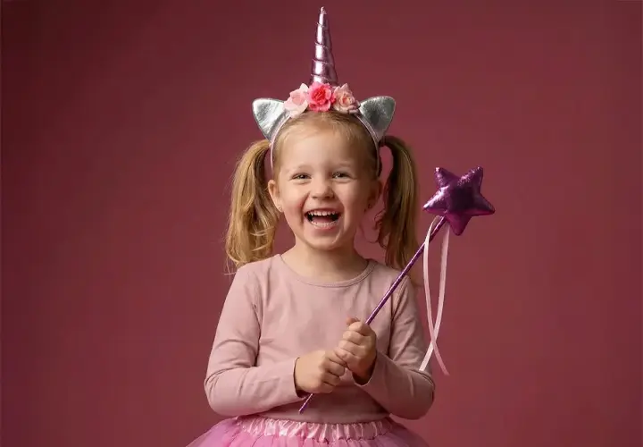 Ein Kind trägt ein Einhorn-Haarband und einen rosa Tüllrock und hält einen lilafarbenen Sternenstab vor einem zartrosa Hintergrund.